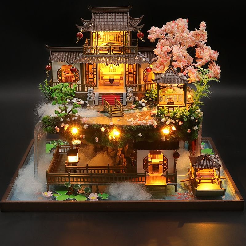 May include: A miniature wooden model of a traditional Chinese house with a cherry blossom tree, a pond, and a small boat. The house has a red roof and white walls, and the pond is surrounded by green plants.
