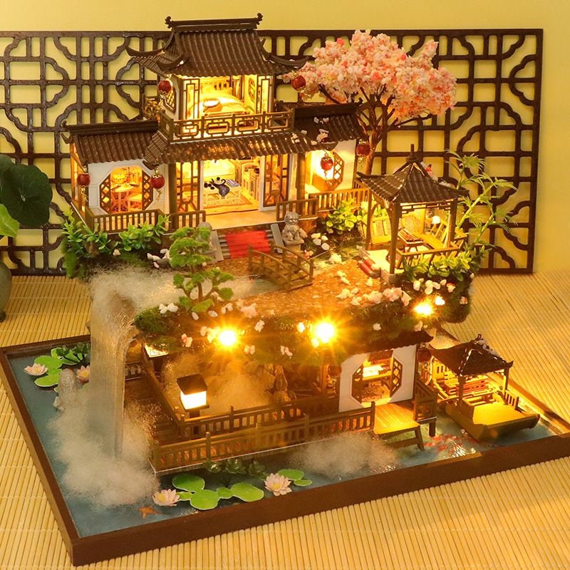 May include: A miniature wooden dollhouse with a traditional Chinese design, featuring a multi-level structure with balconies, a red roof, and a small garden with a cherry blossom tree. The dollhouse is lit with warm white LED lights and has a small pond with lily pads.