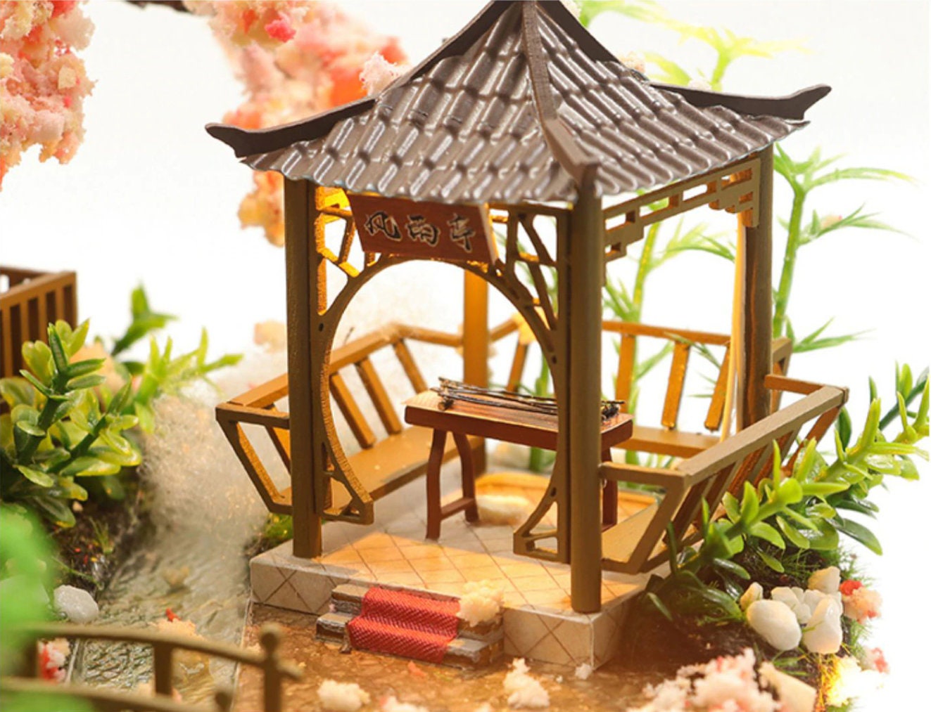 May include: A miniature wooden gazebo with a brown roof and a red rug on the steps. The gazebo has a wooden bench and a table with a small instrument on it. The gazebo is surrounded by green plants and white stones.