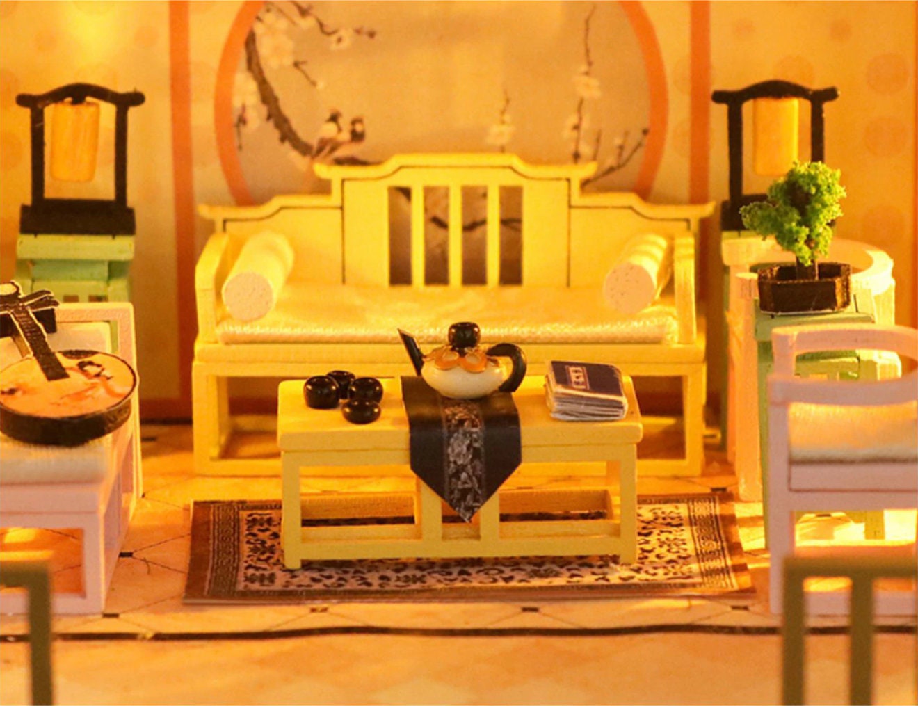 May include: A miniature dollhouse living room with a yellow couch, a yellow coffee table, a teapot, and a book. The room is decorated with a rug and a small potted plant.