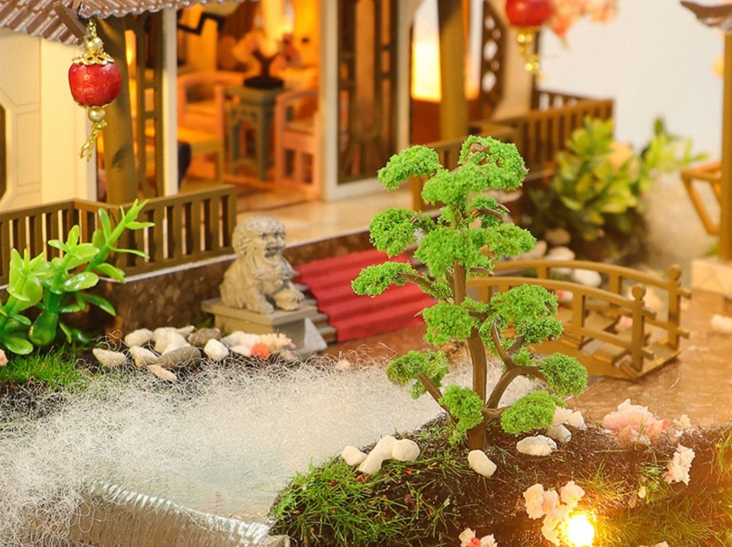 May include: A miniature dollhouse with a red roof and a small green tree in the front yard. The dollhouse has a wooden bridge and a stone lion statue.