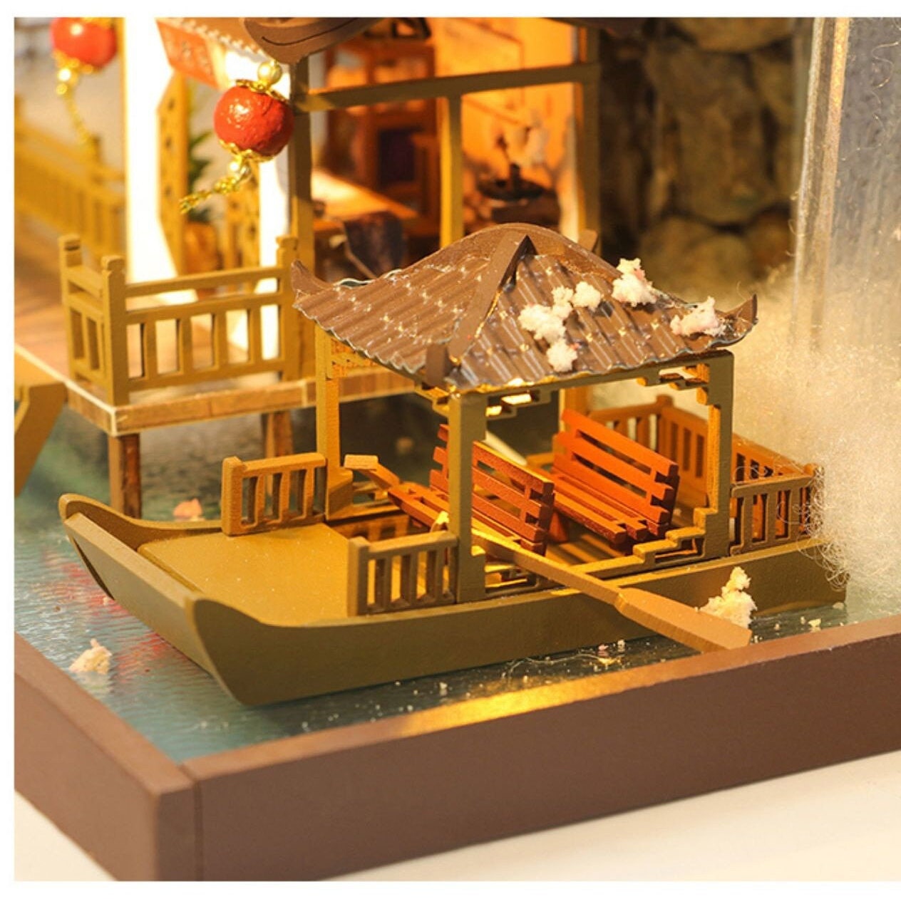 May include: A miniature wooden model of a Chinese-style house with a boat dock and a small boat with a roof and benches. The boat is made of wood and has a brown finish. The house is made of wood and has a white finish. The model is lit from within, creating a warm glow.