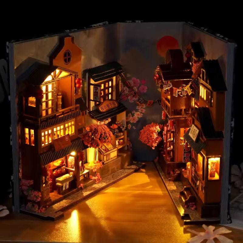 May include: A miniature, illuminated diorama of a Japanese street scene, crafted within the pages of a book. The scene features detailed wooden buildings, glowing windows, and cherry blossom accents, creating a warm, inviting atmosphere.