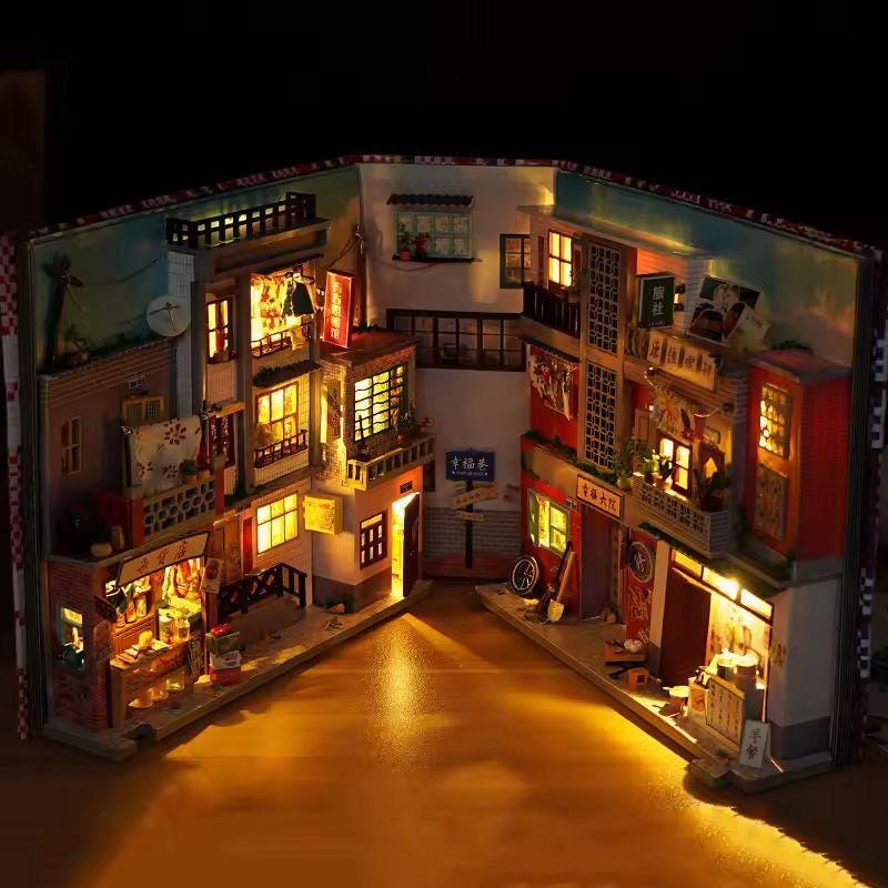 May include: A miniature diorama of a street scene, illuminated with warm lights. The model, built inside a book, features detailed buildings with balconies, shops, and signs. The scene is a charming representation of a small town.