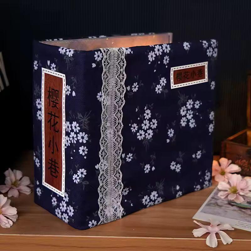 May include: A navy blue photo album with a floral pattern of white flowers. The album has a white lace trim down the center and rectangular labels with Japanese text. The album is sitting on a wooden surface.