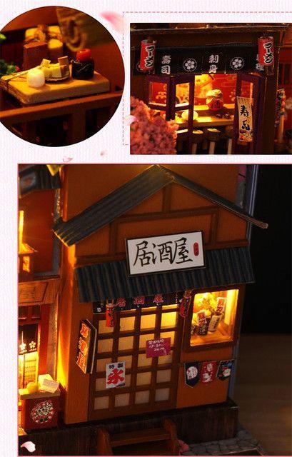 May include: Miniature Japanese restaurant model with a wooden exterior, illuminated windows, and a sign that reads "Izakaya". The model includes small details like paper lanterns and miniature food items.