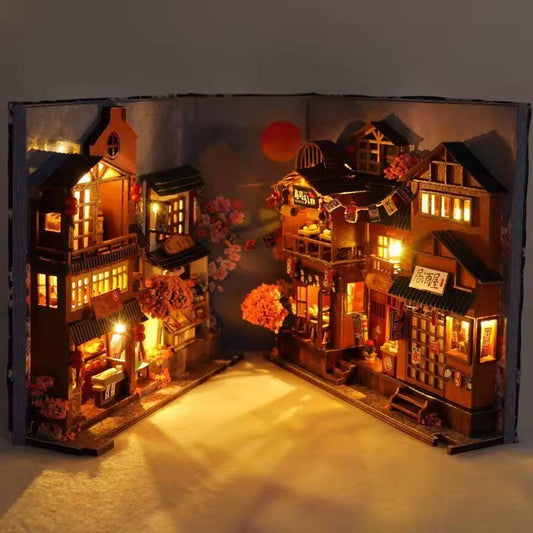 May include: A miniature, illuminated diorama of a Japanese village scene, set within an open book. The detailed wooden structures feature warm interior lighting, a red sun, and cherry blossom accents, creating a cozy, inviting atmosphere.