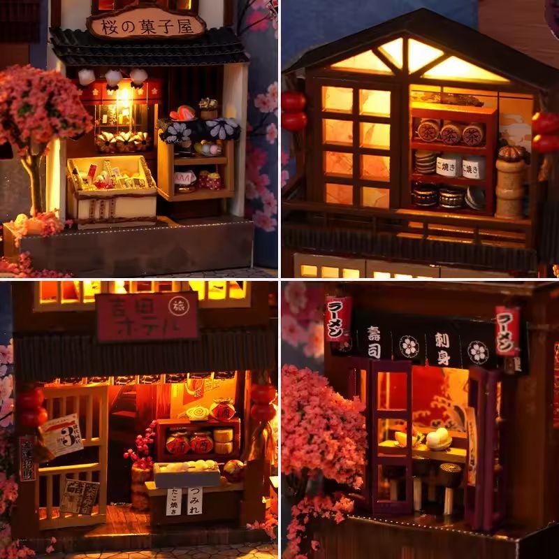 May include: Miniature Japanese shop models with intricate details. The models feature traditional architecture, illuminated interiors, and cherry blossom accents. The shops include a bakery, a ramen shop, and a sushi restaurant.