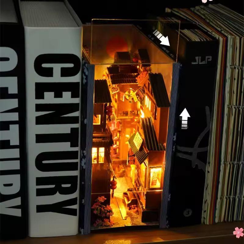 May include: A miniature, illuminated diorama of a Japanese street scene nestled between book covers. The scene features detailed buildings, warm lighting, and a sense of depth. The book spines are white and black, with the word "CENTURY" visible on one.