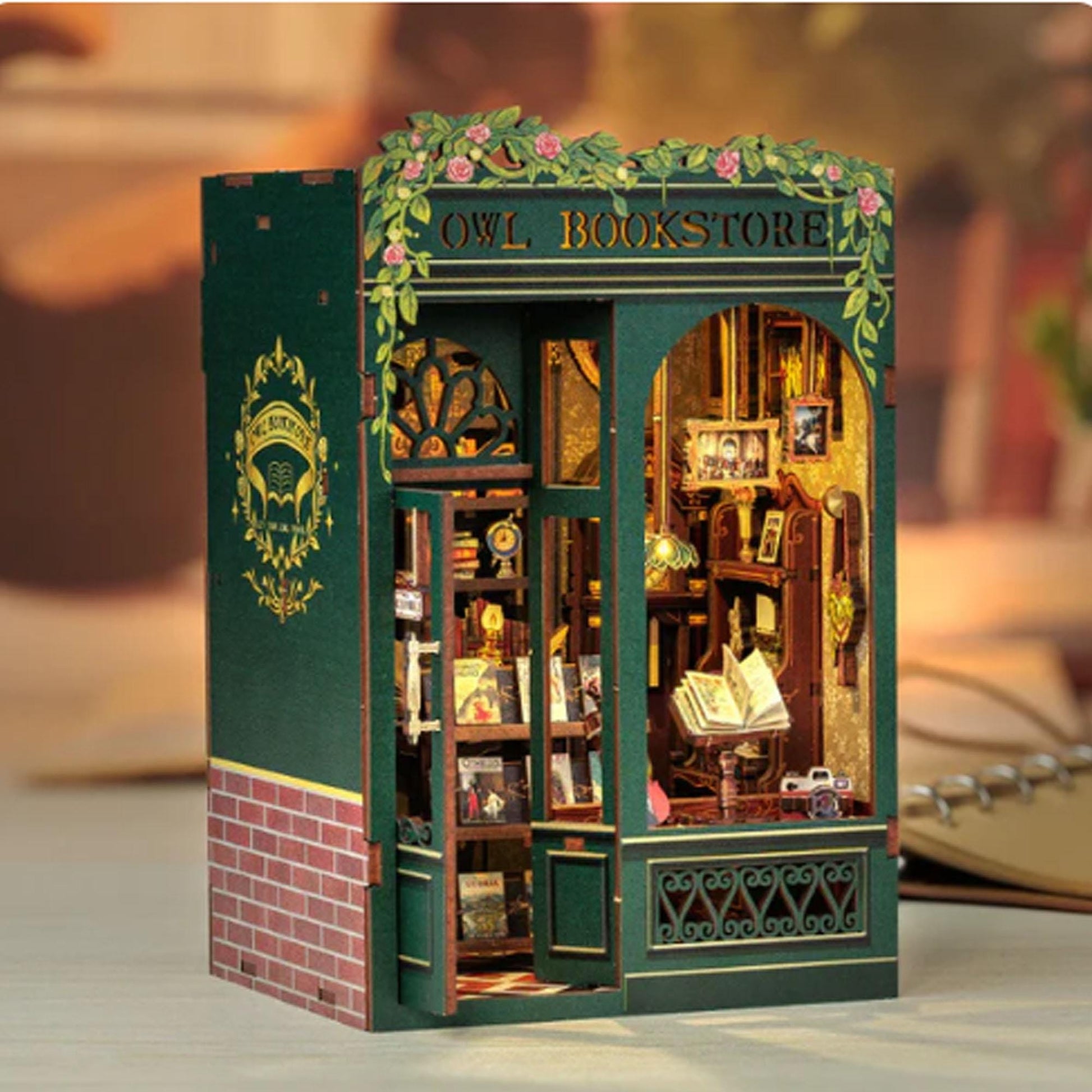 May include: A miniature wooden model of a bookstore with a green exterior and a sign that reads "Owl Bookstore". The storefront has a window with a view of the interior, which includes bookshelves, a desk, and a chair. The model is detailed with ivy vines and flowers.
