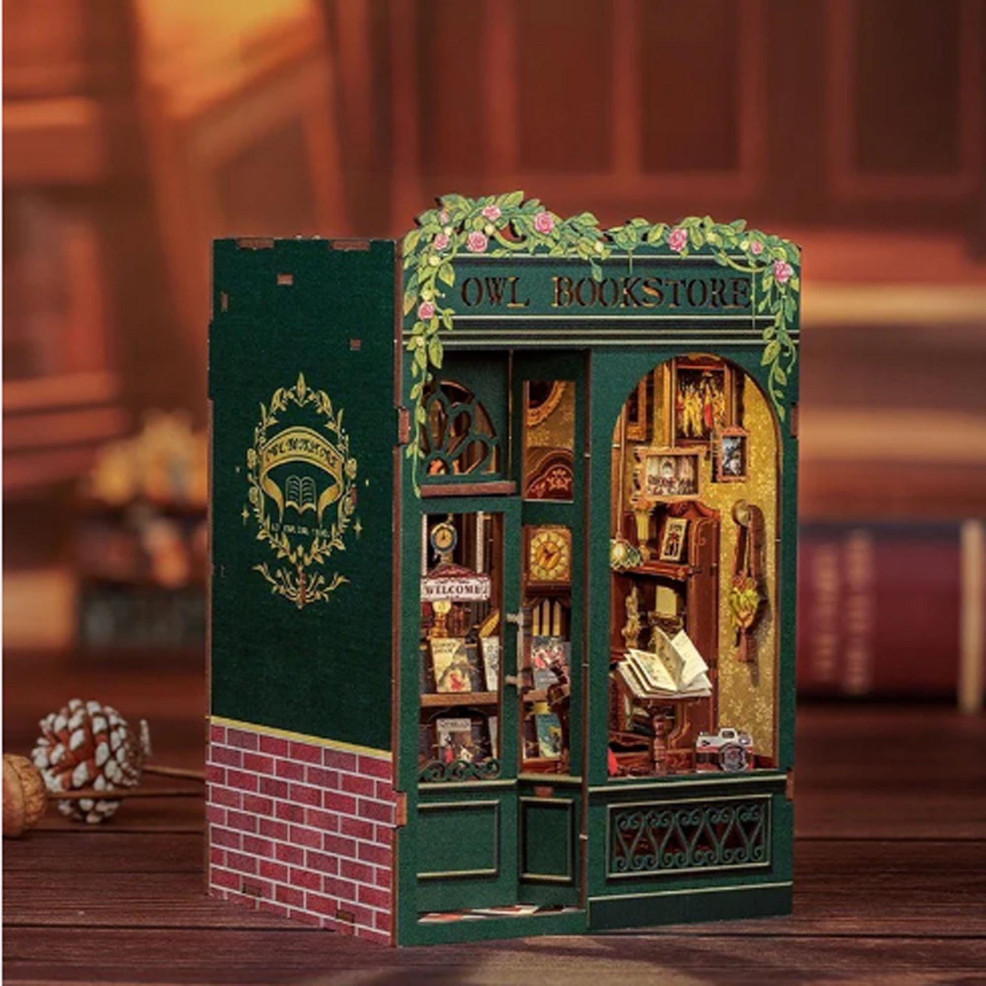 May include: A miniature wooden model of a bookstore with a green exterior and the words "Owl Bookstore" above the entrance. The storefront has a window display with books, a clock, and a welcome sign. The model is detailed with brickwork, vines, and a sign with the words "Owl Bookstore" above the entrance.