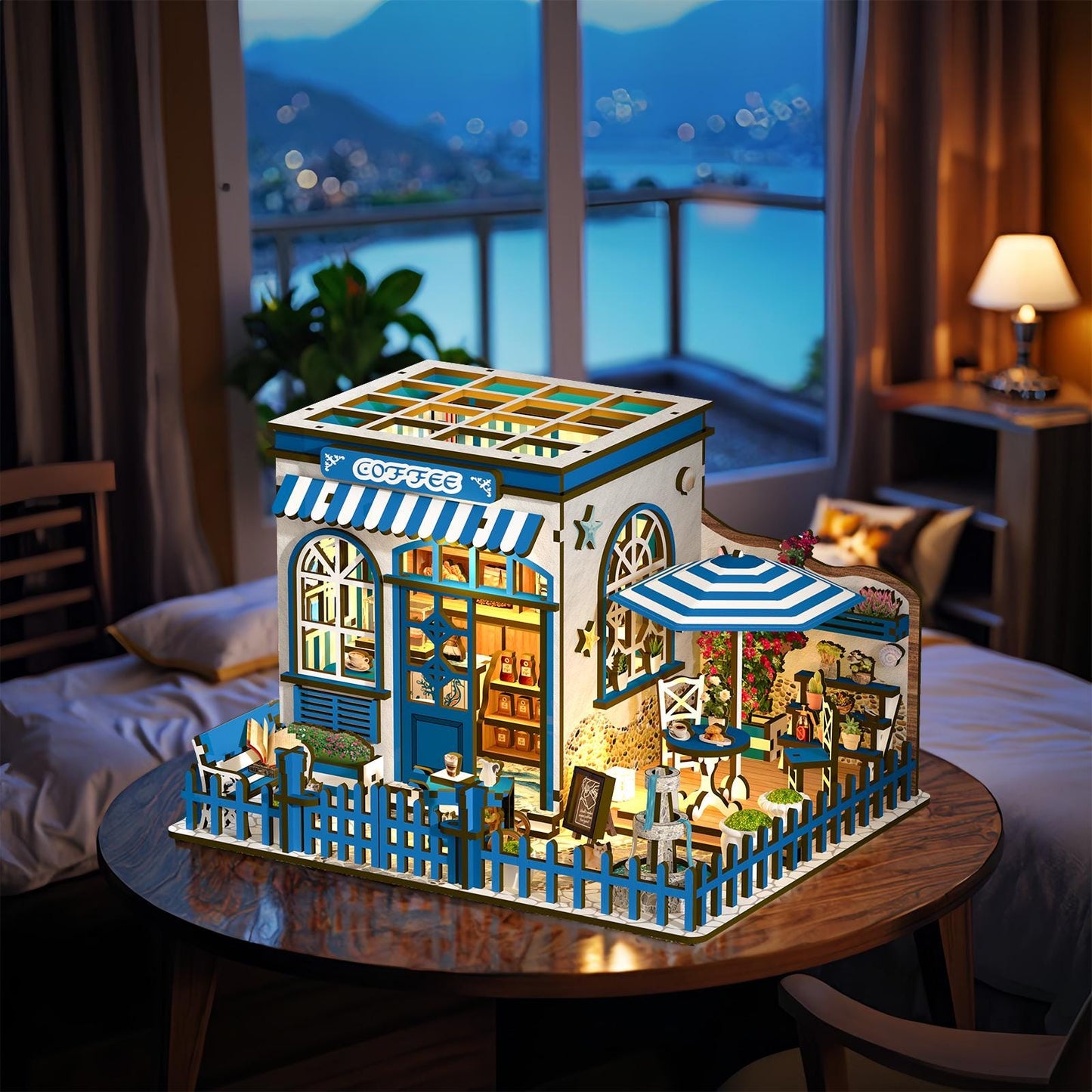 May include: A miniature wooden coffee shop model with blue and white accents. The model features a striped awning, a small outdoor seating area with a blue and white umbrella, and a blue picket fence. The word "COFFEE" is visible on the awning.