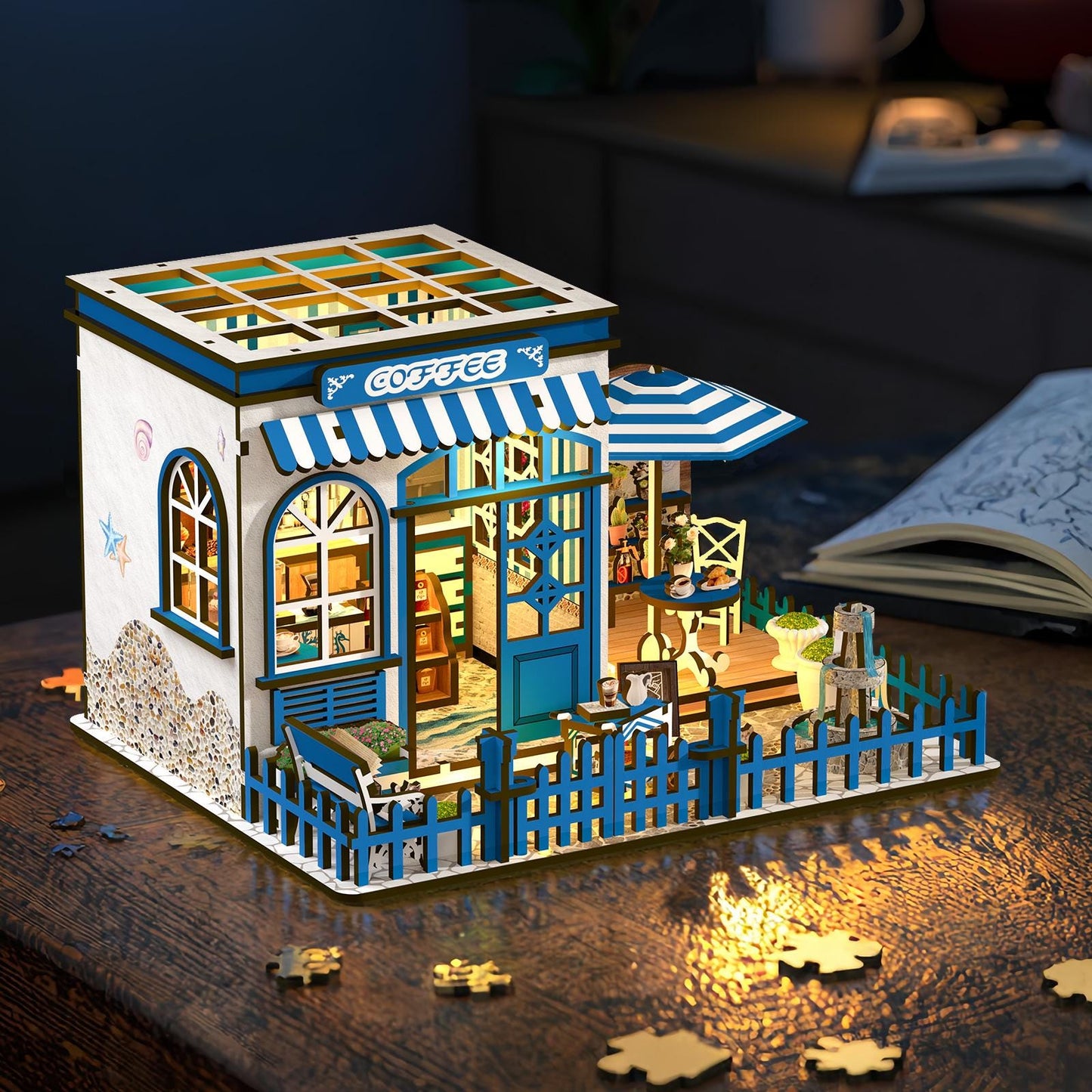 May include: A miniature wooden coffee shop model with blue and white accents. The model features a striped awning, a small fence, and the word "Coffee" above the door. The interior is lit, and the model includes a small table and chairs.