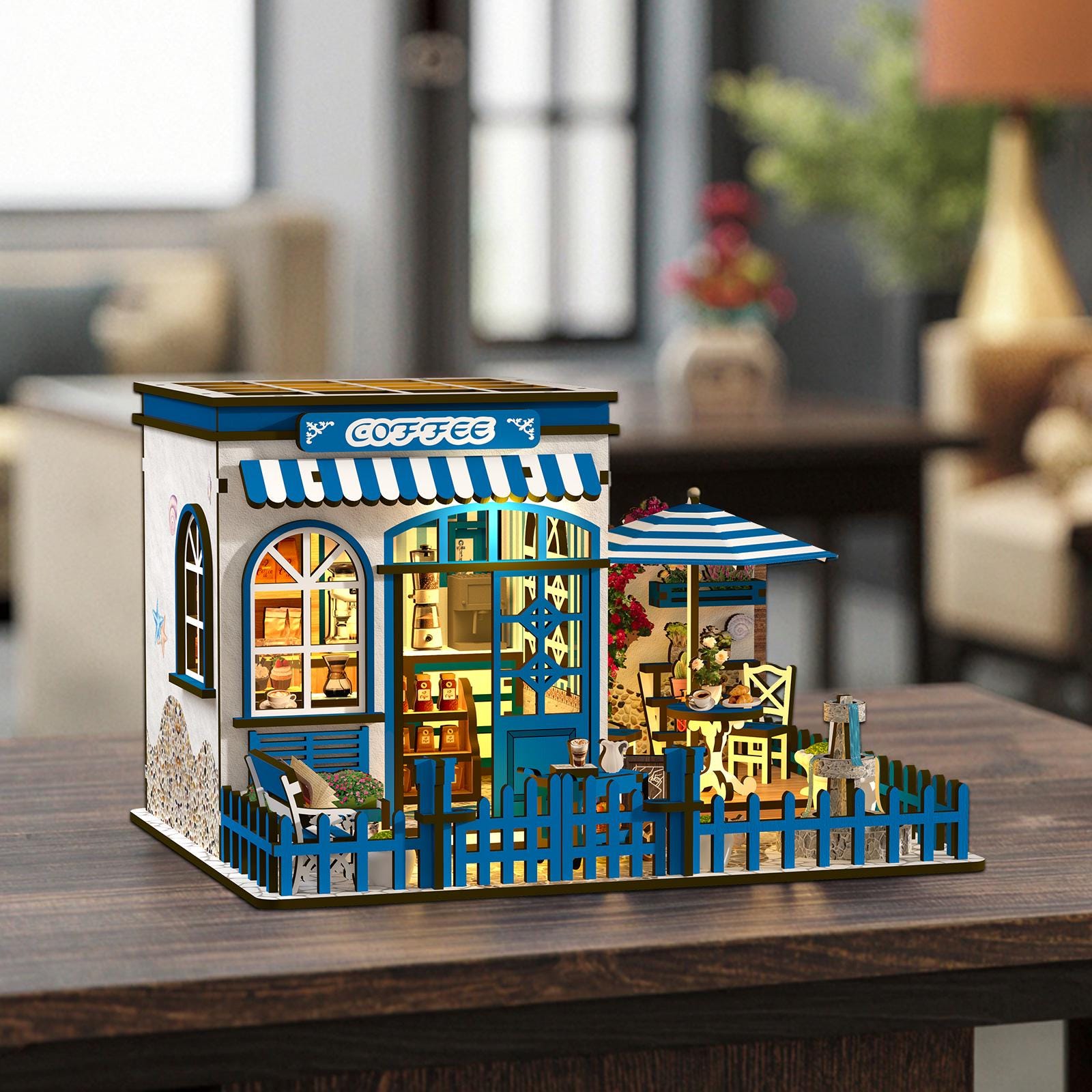May include: A miniature coffee shop model with a blue and white color scheme. The model features a blue awning with the word "COFFEE", a blue picket fence, and a small outdoor seating area with a striped umbrella. The shop is illuminated with warm lighting.