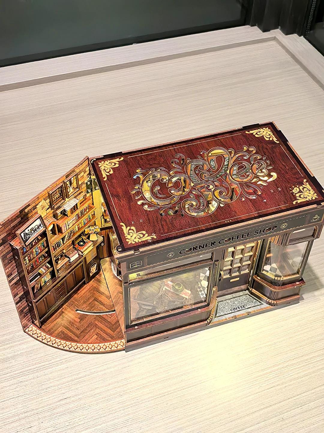 May include: A detailed, three-dimensional model of a Corner Coffee Shop. The model features a brown facade with decorative gold accents, a glass display window, and a detailed interior with miniature furniture and books. The roof has an ornate design.