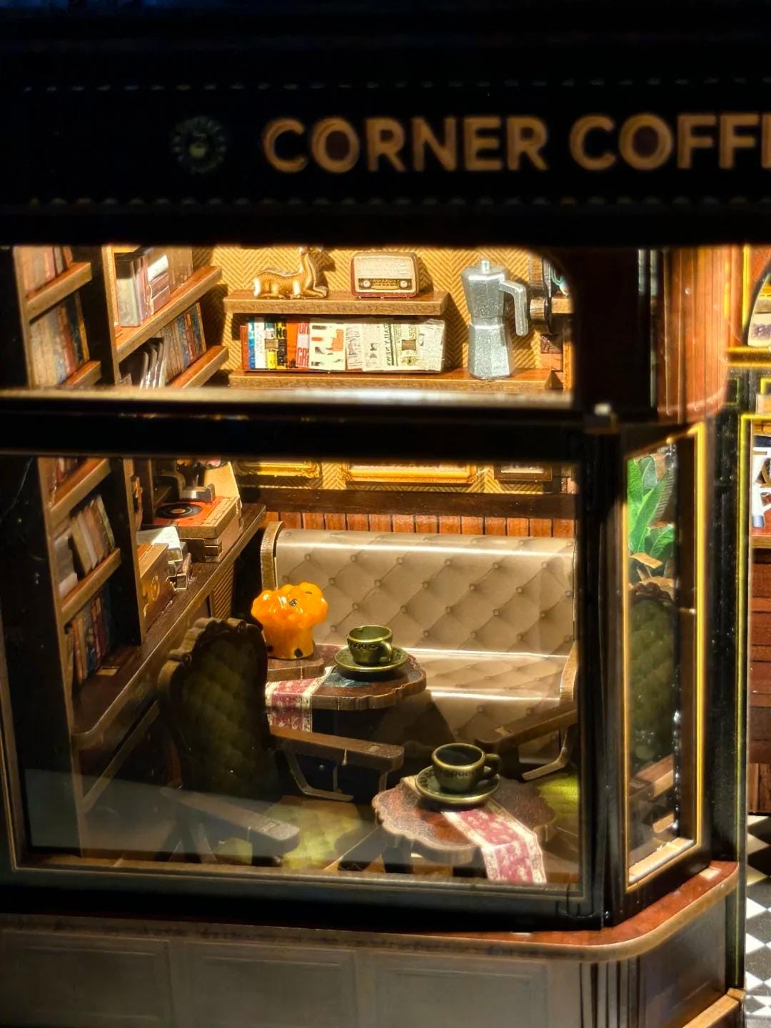 May include: Miniature corner coffee shop model with the text "CORNER COFFEE" above the window. The interior features bookshelves, a sofa, chairs, tables with coffee cups, and a coffee maker. The shop is lit with warm lighting.