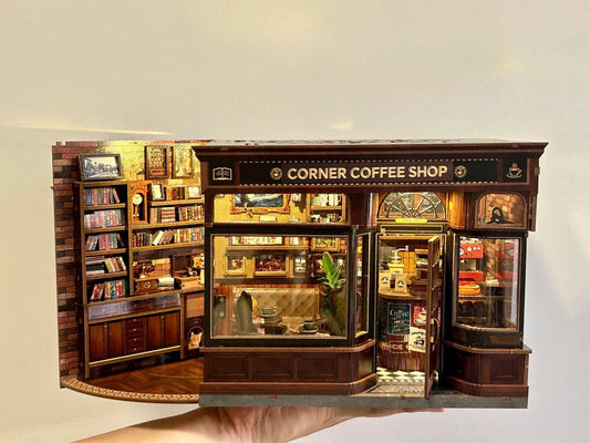 May include: Miniature Corner Coffee Shop model with detailed interior and exterior. The shop features a brick facade, wooden trim, and a sign that reads "CORNER COFFEE SHOP". Inside, there are shelves, books, and miniature furniture. The shop is lit with small lights.