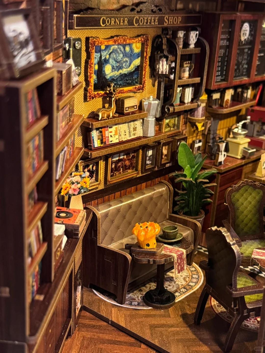 May include: Miniature model of a coffee shop interior. The scene includes bookshelves, a sofa, tables, and framed artwork, including a painting of Van Gogh's "Starry Night." The shop is decorated with small details like a coffee maker and a radio.