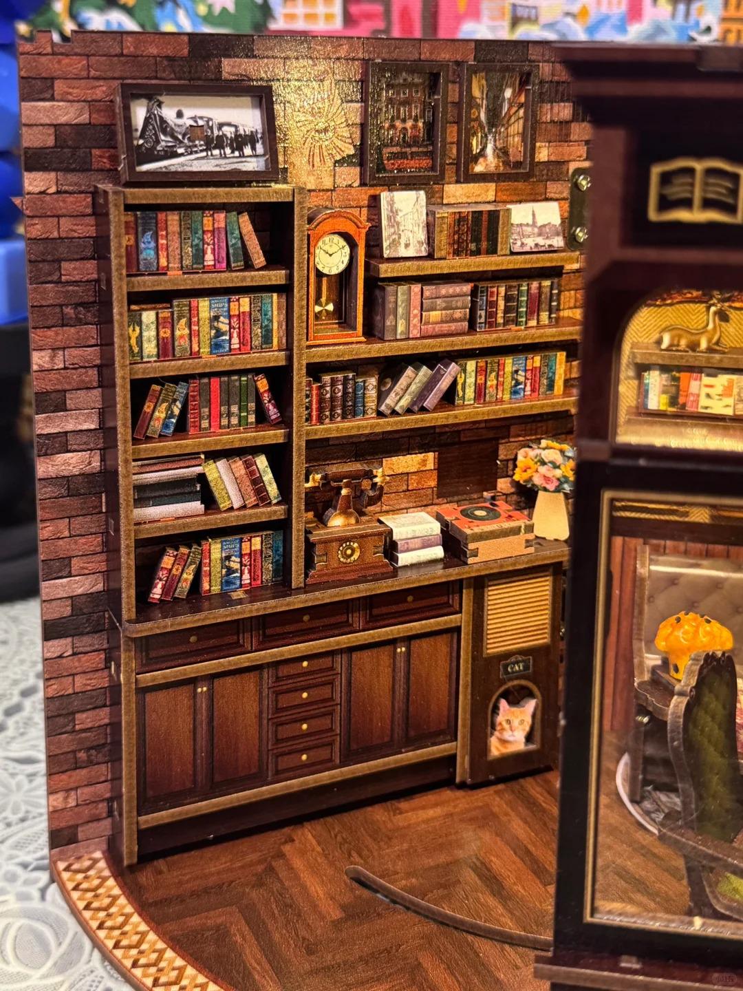 May include: A miniature wooden library scene with a brick wall backdrop. The scene includes bookshelves filled with tiny books, a grandfather clock, framed pictures, and a small cat house. The overall color scheme is warm, with browns and reds.