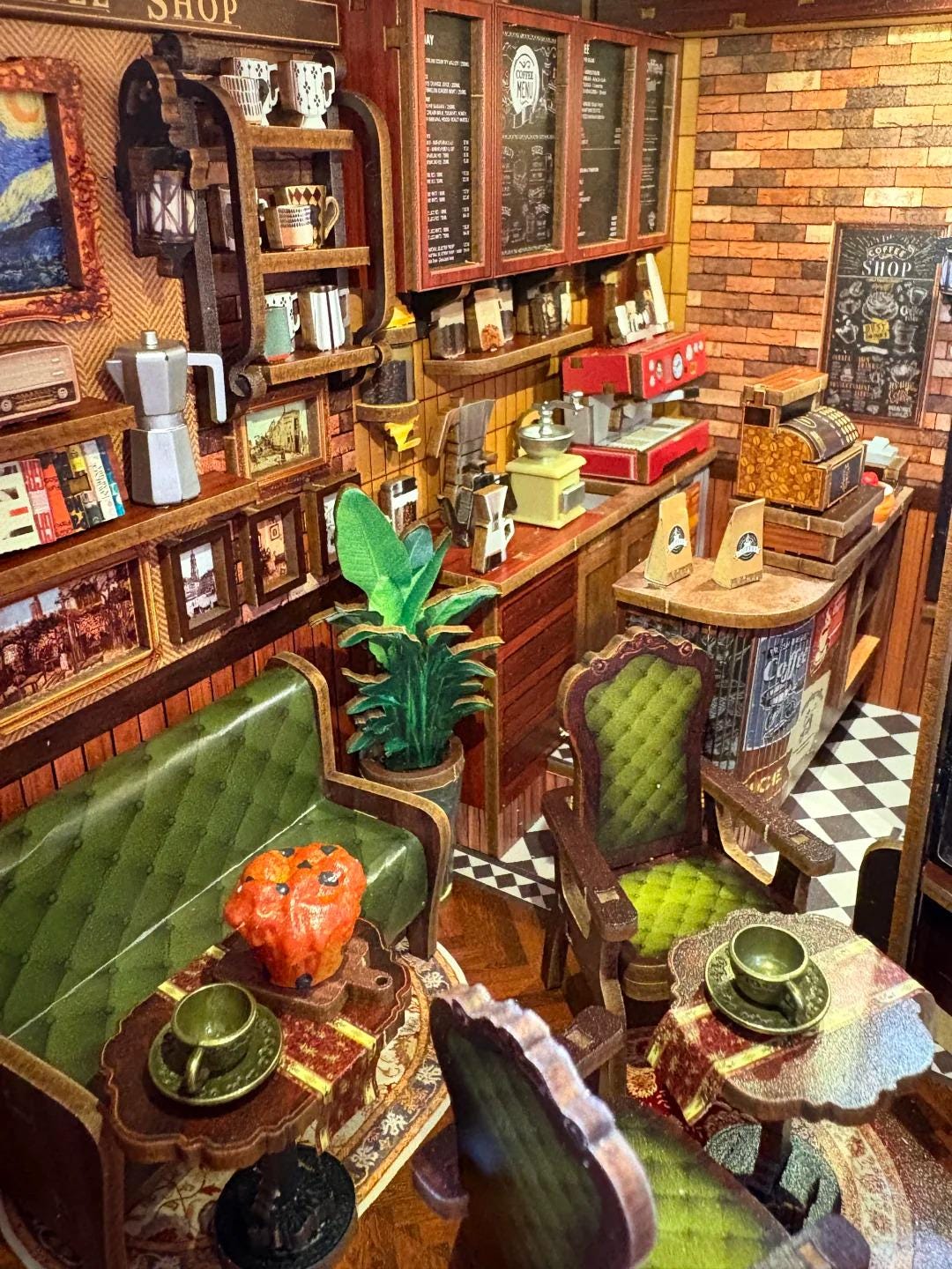 May include: A miniature coffee shop diorama with detailed interior elements. Features include a green sofa, wooden tables, chairs, and a counter with a cash register. The walls are decorated with framed art and shelves filled with coffee-related items.