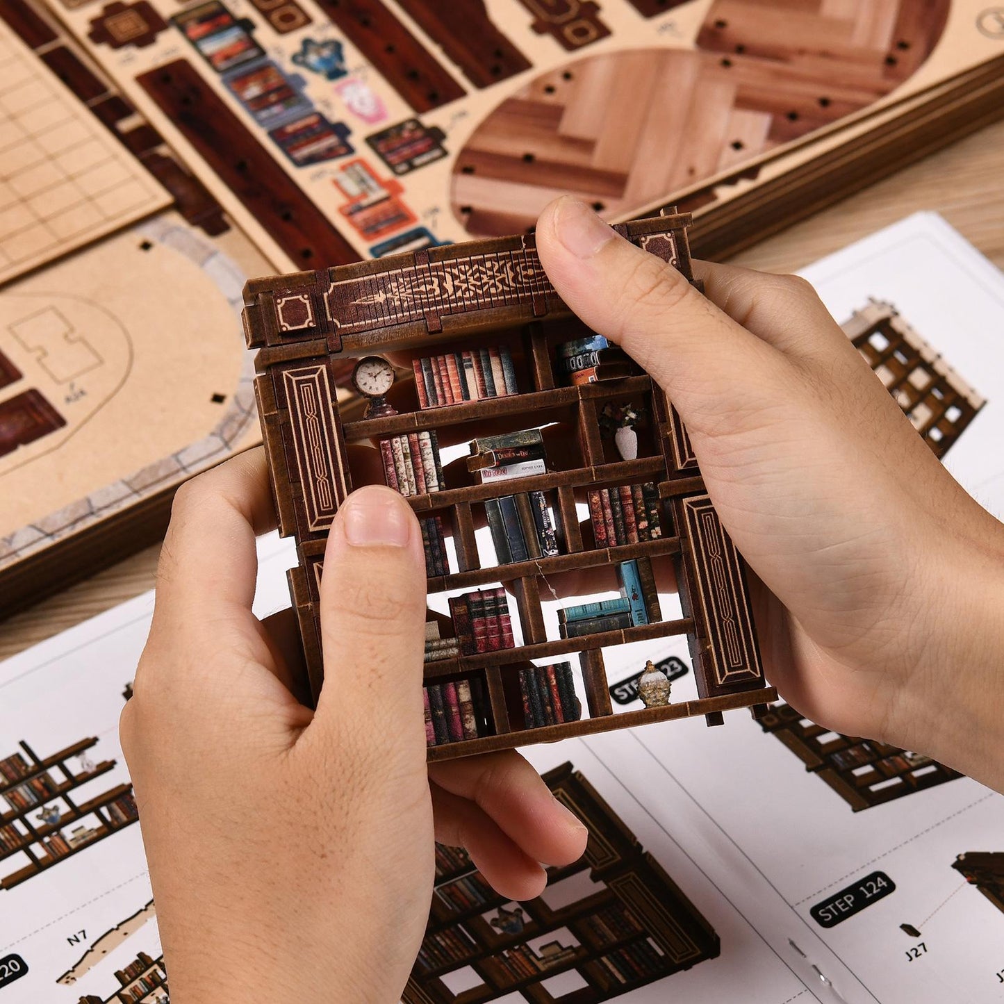 May include: A miniature wooden bookshelf puzzle, featuring detailed shelves filled with tiny books, a clock, and decorative elements. The puzzle is held in hands, with assembly instructions visible in the background. The wood has a dark brown finish.