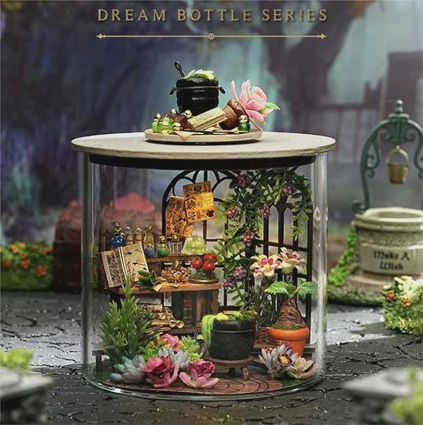 May include: A miniature diorama inside a clear glass cylinder, featuring a fantasy scene with a small cauldron, flowers, and a well. The words "DREAM BOTTLE SERIES" are at the top. The scene includes miniature books, plants, and a small table.