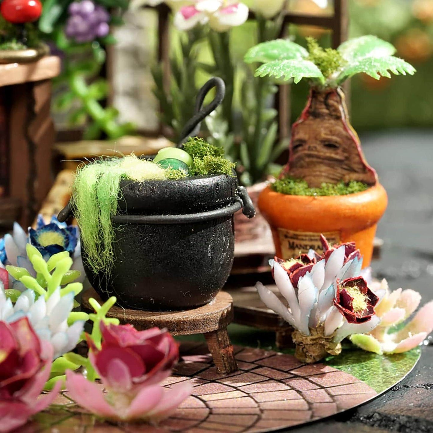 May include: Miniature black cauldron with green and yellow contents, set on a small wooden stool. The scene includes a small orange pot with a tree-like plant, and colorful artificial flowers. The setting is a miniature garden.