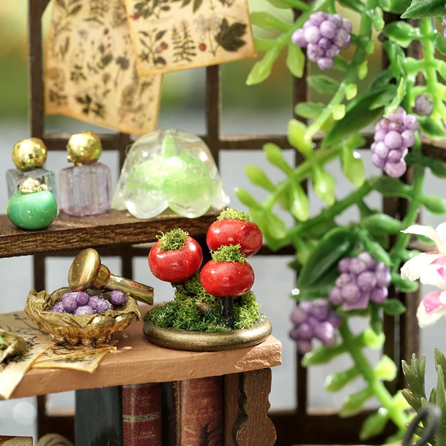 May include: Miniature scene featuring a shelf with tiny bottles, red mushroom-like objects, and a small bowl of purple grapes. The scene includes a green dome and decorative floral elements, creating a whimsical, fairy-tale aesthetic.
