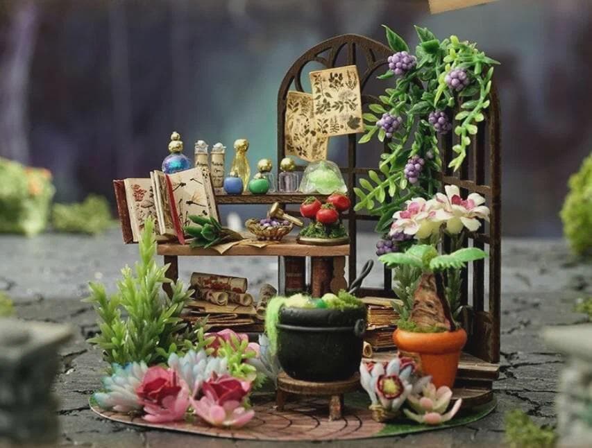 May include: Miniature fairy garden scene with a wooden shelf, books, and potion bottles. Features a black cauldron, potted plants, and a decorative arch with flowers and grapes. The scene is set on a stone surface.