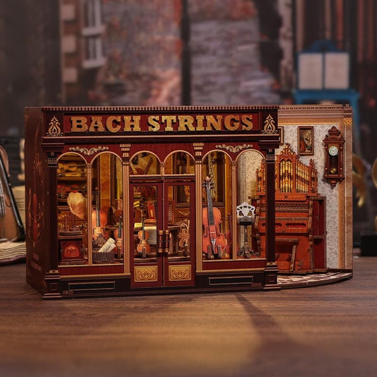 May include: A detailed miniature model of a music shop, featuring the text "BACH STRINGS" above the entrance. The shop's facade is a deep red with gold accents, showcasing violins and other instruments. The model includes an adjacent room with an organ and clock.